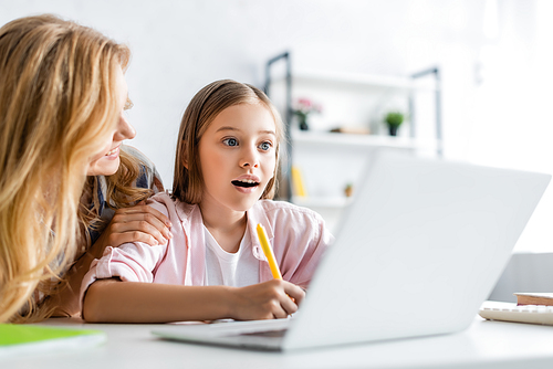 Selective focus of smiling woman embracing daughter writing on notebook during online education at home