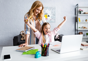 Selective focus of smiling mother looking at cheerful kid showing yeah gesture during online education