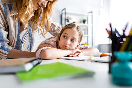 Selective focus of mother embracing sad daughter near stationery on table