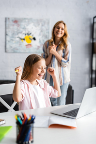 Selective focus of smiling child showing yeah gesture during online education near positive mother at home