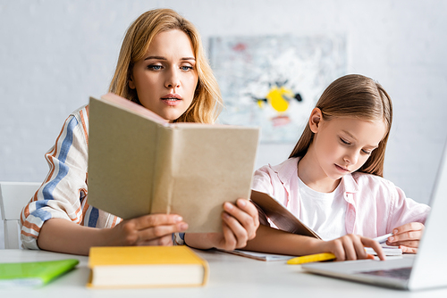 Selective focus of woman reading book while helping daughter during online education at home