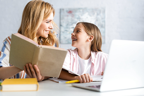 Selective focus of cheerful child looking at mother with book near laptop at table