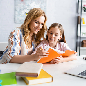 Selective focus of smiling mother and kid looking at notebook near laptop and books on table
