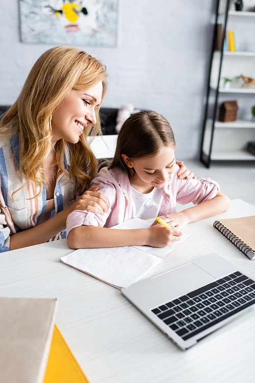 Selective focus of smiling woman embracing kid writing on notebook near laptop on table