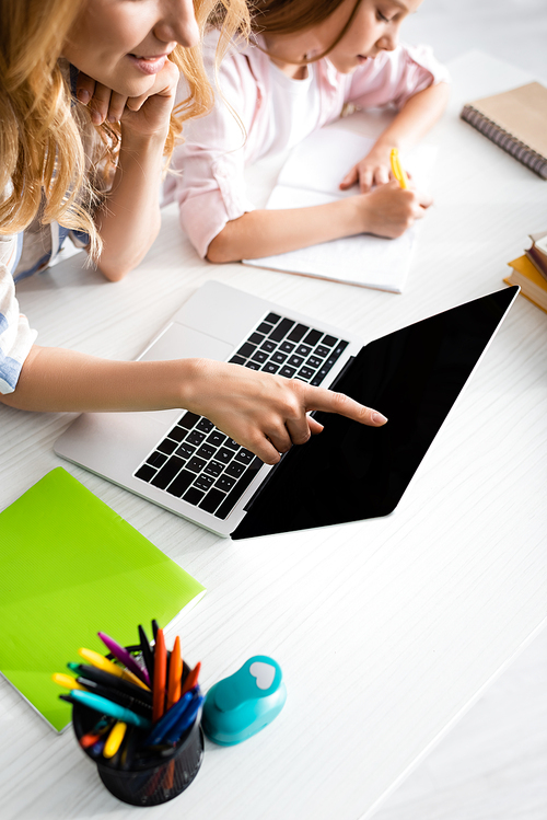 High angle view of woman pointing with finger at laptop while daughter writing on notebook at table