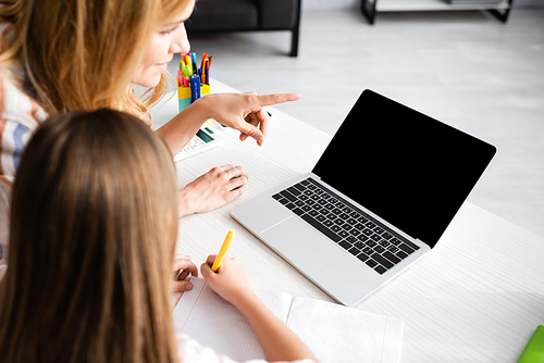 Selective focus of woman pointing with finger at laptop near kid writing on notebook during electronic learning at home