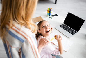 Selective focus of positive kid looking at mother while holding pen near notebook and laptop on table