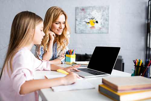 Selective focus of smiling woman standing near kid using laptop during electronic learning at home