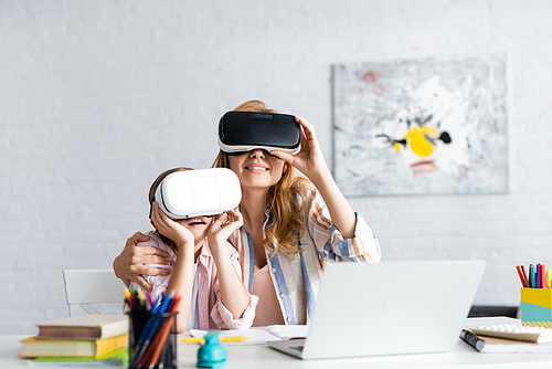 Selective focus of smiling woman embracing daughter while using virtual reality headsets near laptop and books