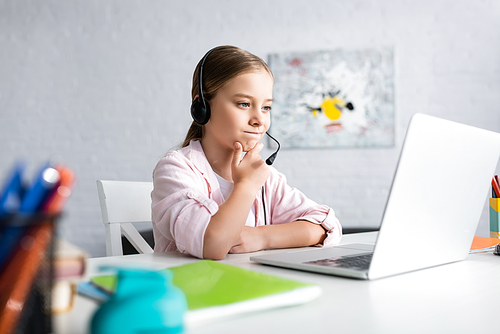 Selective focus of thoughtful kid in headset using laptop near stationery on table