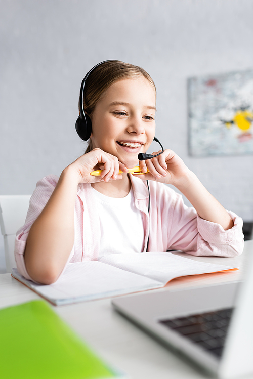 Selective focus of smiling child in headset holding pen and looking at laptop on table
