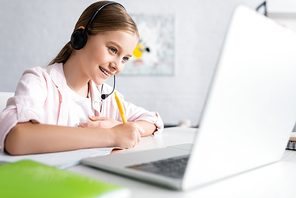 Selective focus of positive kid in headset writing on notebook and using laptop at home