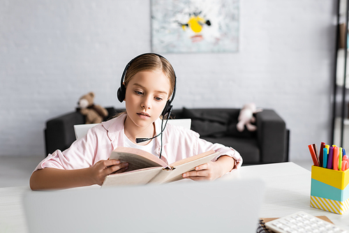Selective focus of child in headset reading book near laptop on table