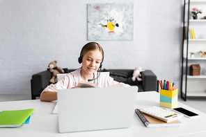 Selective focus of smiling kid in headset reading book near gadgets on table