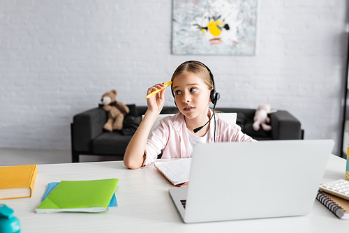 Selective focus of pensive child holding pen while using headset near laptop and stationery on table