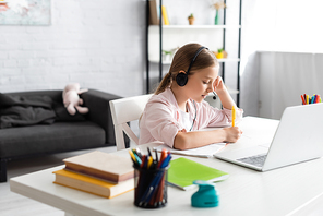 Selective focus of child writing on notebook and using headset near laptop on table