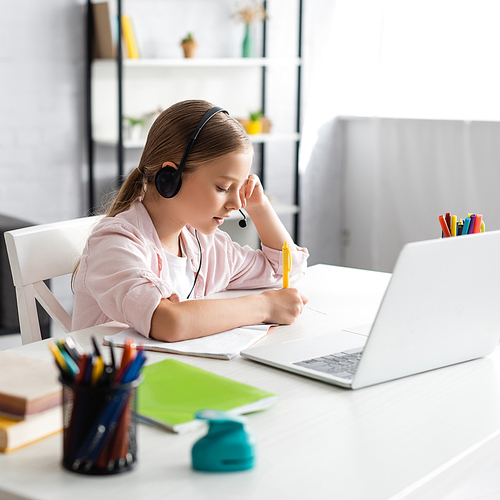 Selective focus of kid in headset writing on notebook near laptop and stationery on table