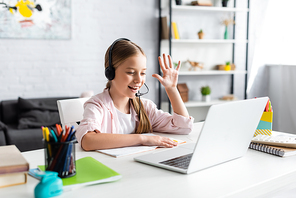 Selective focus of positive kid in headset waving hand during webinar at home
