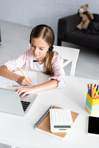 Selective focus of kid writing on notebook while using headset and laptop on table