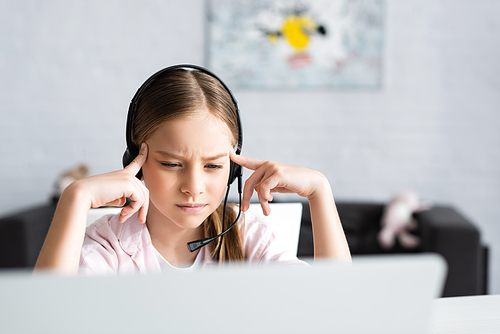 Selective focus of thoughtful kid in headset looking at laptop at home