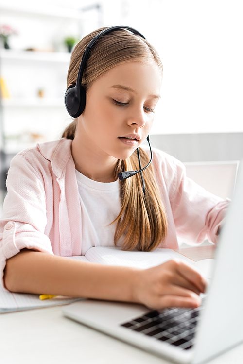 Selective focus of cute child in headset typing on laptop near notebook on table