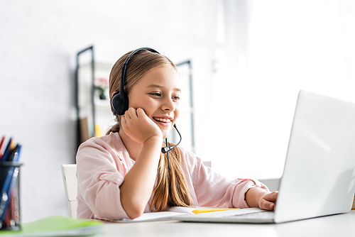 Selective focus of smiling kid using headset and laptop at table
