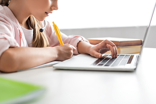 Cropped view of kid in headset writing on notebook and using laptop at home