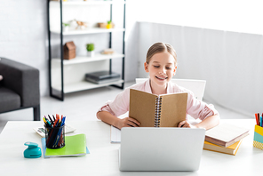 Selective focus of smiling kid holding notebook near laptop and books on table at home
