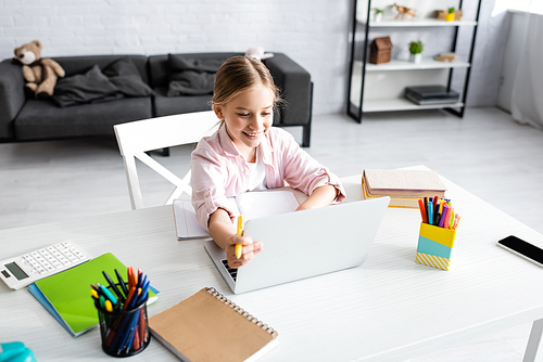 Selective focus of smiling child holding pen and using laptop near books on table