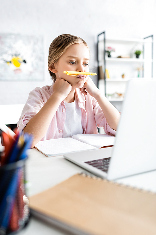 Selective focus of kid holding pen near lips during electronic learning at home