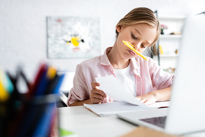 Selective focus of kid with pen near mouth looking at notebook near laptop