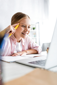 Selective focus of smiling child holding pen and looking at laptop on table