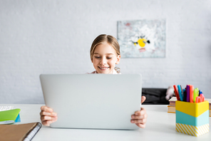 Selective focus of smiling child holding laptop near books on table