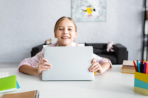 Selective focus of smiling kid holding laptop and  near stationery