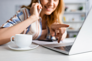 Cropped view of smiling woman pointing with finger at laptop while talking on smartphone near cup of coffee on table