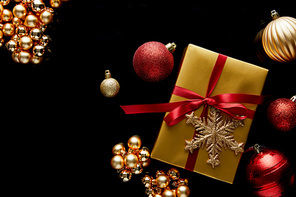 top view of shiny golden Christmas gift with red ribbon and snowflake near baubles isolated on black