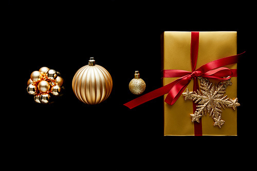 flat lay with shiny golden Christmas gift with red ribbon and snowflake near baubles isolated on black