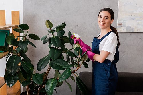 Side view of smiling cleaner holding spray bottle near plant at home