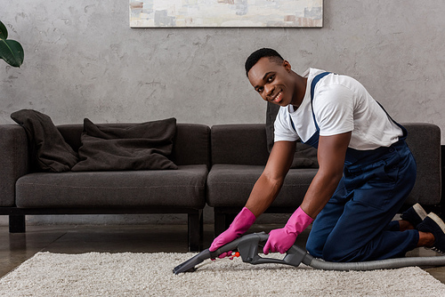 African american cleaner in uniform smiling at camera while cleaning carpet at home