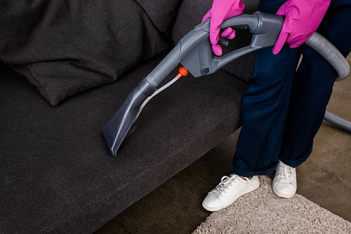 Cropped view of cleaner in rubber gloves holding vacuum cleaner near couch