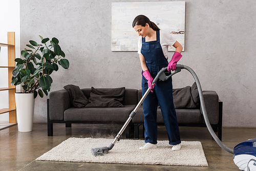 Beautiful cleaner in workwear and rubber gloves using vacuum cleaner with hot steam on carpet in living room