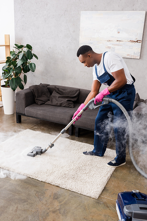 African american cleaner vacuuming carpet with hot steam in living room