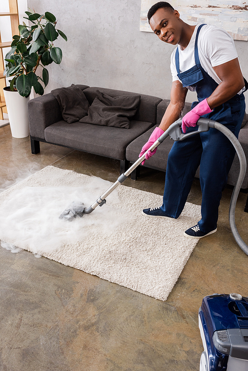 African american cleaner in uniform and rubber gloves smiling at camera while cleaning carpet with hot steam