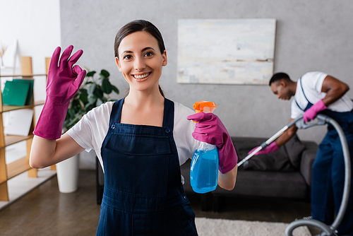 Selective focus of smiling cleaner showing ok gesture and holding detergent near african american colleague with vacuum cleaner at home