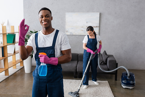 Selective focus of smiling african american cleaner showing okay and holding detergent near colleague using vacuum cleaner