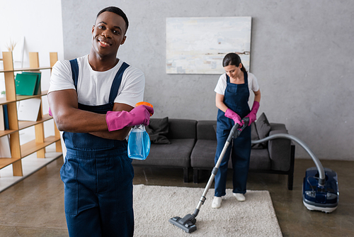 Selective focus of african american cleaner holding detergent and smiling at camera near colleague cleaning carpet