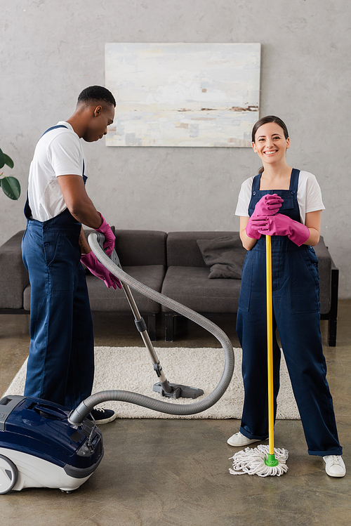 Smiling cleaner holding mop near african american colleague cleaning carpet at home