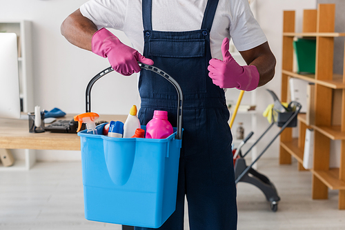 Cropped view of African american cleaner showing thumb up and holding bucket of detergents in office