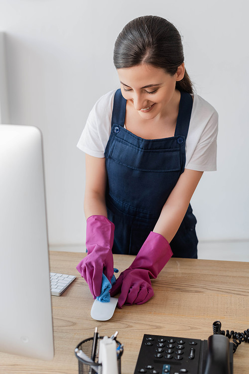 Selective focus of smiling cleaner holding rag while cleaning computer mouse on office table