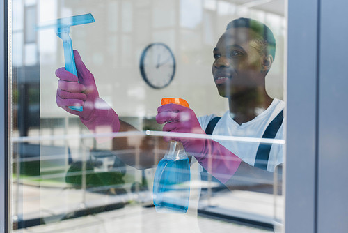 Selective focus of african american cleaner in rubber gloves using detergent and squeegee handle while cleaning window